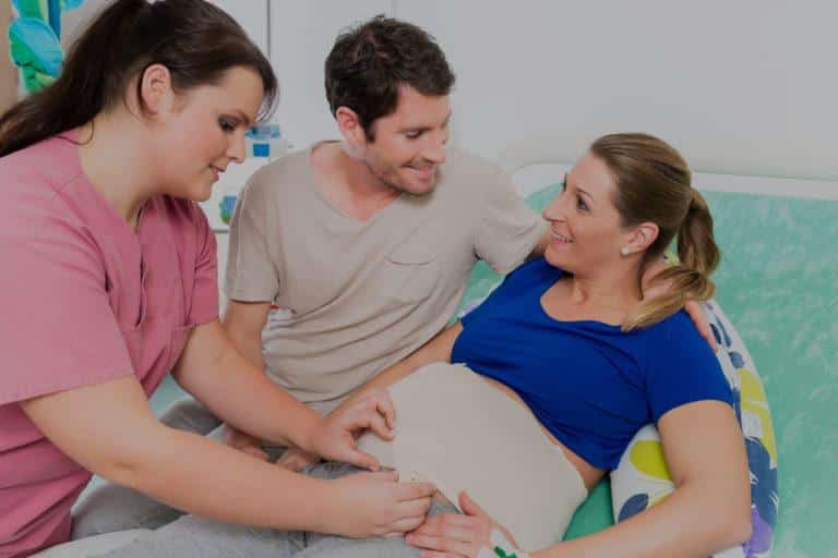 episiotomie Femme souriante accompagné de son mari, allongée sur un lit et se faisant soigner par une infirmière